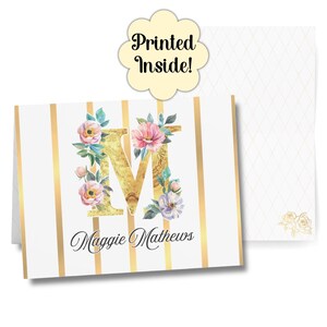 Puede incluir: Un juego de papelería personalizado con una letra "M" dorada adornada con flores de acuarela. La tarjeta tiene rayas verticales doradas y el nombre "Maggie Mathews" en una elegante escritura. El sobre tiene un diseño floral. El texto "Printed Inside!" también es visible.