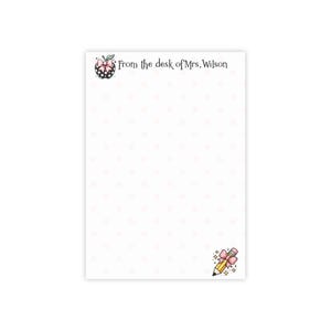 May include: White notepad with a pink heart pattern. The top of the notepad has a black and white apple illustration with a pink bow and the text "From the desk of Mrs. Wilson". A pencil illustration with a pink bow is at the bottom.