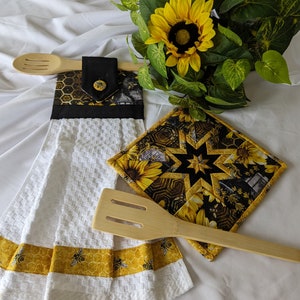 May include: A white kitchen towel with a yellow and black honeycomb pattern border. A matching yellow and black pot holder with a star design is on the towel. A wooden spoon and spatula are on the towel.
