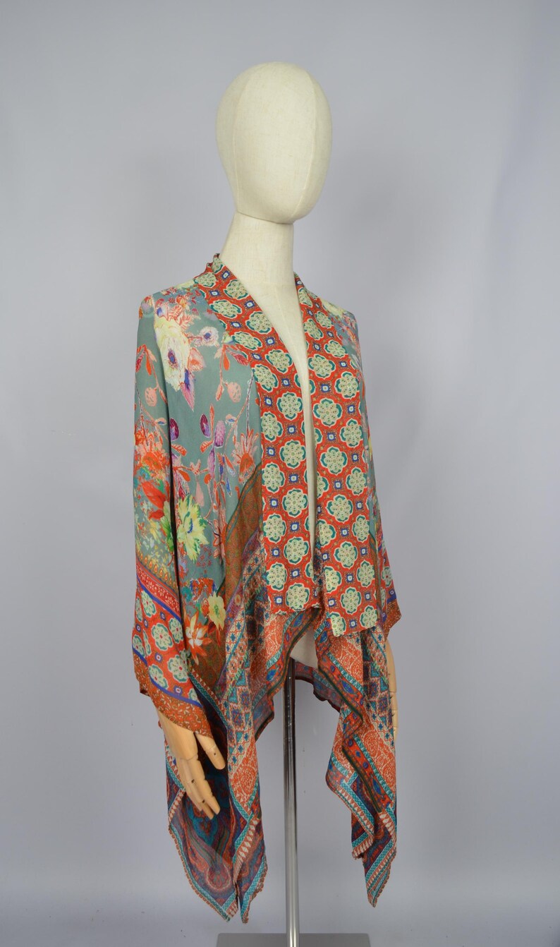 May include: A patterned kimono-style garment with a floral and geometric design in shades of teal, orange, red, and blue. The fabric drapes over a mannequin, showcasing the flowing design and vibrant colours. The garment is a stylish addition to any wardrobe.
