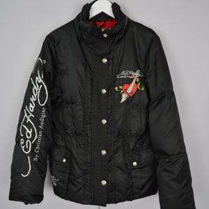 Puede incluir: Chaqueta acolchada negra con cierre de cremallera y detalles de botones. La chaqueta presenta un logotipo bordado en blanco que dice "Ed Hardy by Christian Audigier" en la manga izquierda y un corazón rojo con una daga y detalles florales en la parte delantera.