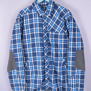 May include: Blue and white plaid button-up shirt with gray elbow patches. The shirt has a classic collar, buttoned cuffs, and two front pockets. The shirt is hanging on a hanger against a light purple background.