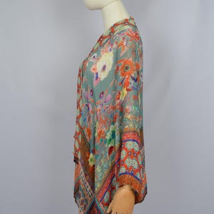 May include: A patterned top in teal, orange, and red, shown on a mannequin. The top has a flowing, asymmetrical cut with floral and paisley designs. The fabric looks lightweight and semi-transparent. The top is a piece of clothing.