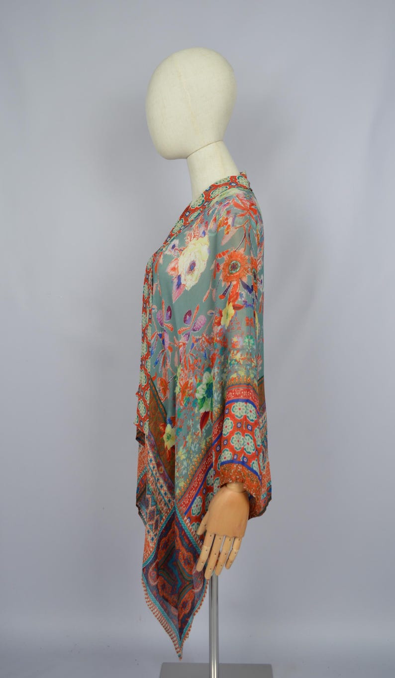 May include: A patterned top in teal, orange, and red, shown on a mannequin. The top has a flowing, asymmetrical cut with floral and paisley designs. The fabric looks lightweight and semi-transparent. The top is a piece of clothing.