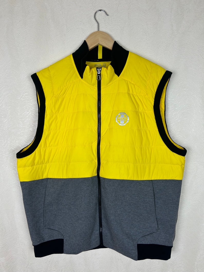 Puede incluir: Un chaleco acolchado sin mangas amarillo y gris con una cremallera negra y ribetes negros. El chaleco tiene un logotipo en la parte delantera que dice "Polo" y presenta un peque&ntilde;o logotipo con un caballo y un jinete.