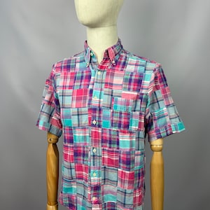 Può includere: Una camicia a maniche corte con bottoni e un motivo a quadri patchwork in tonalità rosa, blu e viola.