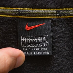 May include: Black Nike tag with a red swoosh logo and white text. The tag says "SIZE L HEIGHT 183", "GB 42/44 1 52/55", "F 52/56 0 52/56", "MADE IN LAOS P.O.R.", and "BRIQUE A LAOS P.D.R.".