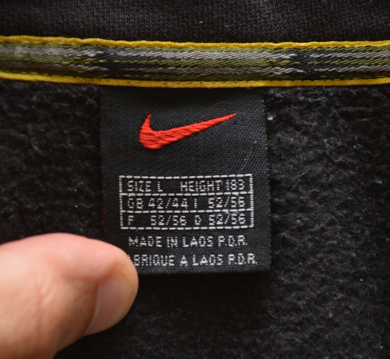 May include: Black Nike tag with a red swoosh logo and white text. The tag says "SIZE L HEIGHT 183", "GB 42/44 1 52/55", "F 52/56 0 52/56", "MADE IN LAOS P.O.R.", and "BRIQUE A LAOS P.D.R.".
