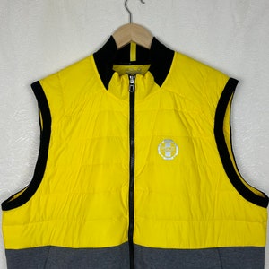Puede incluir: Un chaleco acolchado sin mangas amarillo y gris con cremallera negra y ribetes negros. El chaleco tiene un logotipo en la parte delantera que dice "Polo Ralph Lauren".