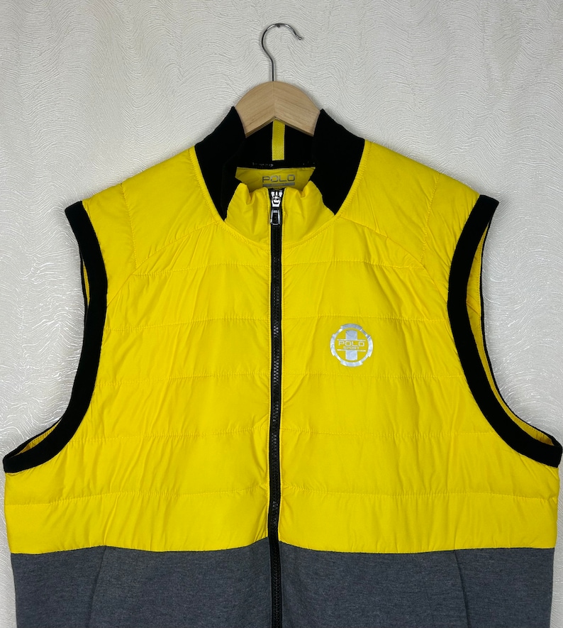 Puede incluir: Un chaleco acolchado sin mangas amarillo y gris con cremallera negra y ribetes negros. El chaleco tiene un logotipo en la parte delantera que dice "Polo Ralph Lauren".