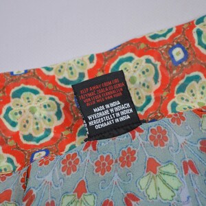 May include: Close-up of a patterned fabric with a tag. The fabric features a red and teal floral design. The tag reads "KEEP AWAY FROM FIRE" in multiple languages, and "MADE IN INDIA".