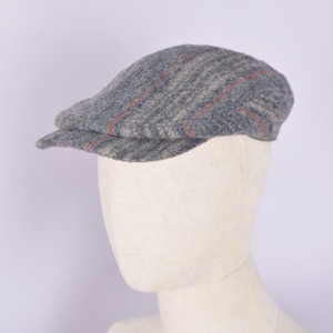 May include: A gray tweed flat cap with red and blue stripes. The hat is displayed on a mannequin head. The cap has a rounded crown and a short, stiff brim. This classic style is suitable for various occasions.