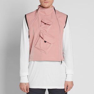 Pode incluir: Um colete rosa sem mangas com vários bolsos e um zíper preto. O colete tem uma gola alta e uma borda preta ao redor das bordas.