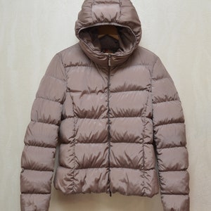 Puede incluir: Una chaqueta acolchada con capucha de color marrón con cierre de cremallera. La chaqueta tiene un diseño acolchado y está hecha de un material ligero.