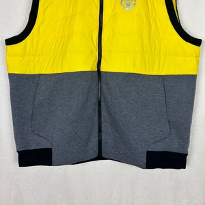 Puede incluir: Un chaleco acolchado amarillo y gris con una cremallera negra y ribetes negros. El chaleco tiene un logotipo en la parte superior izquierda que dice "Polo".
