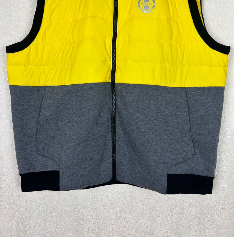 Puede incluir: Un chaleco acolchado amarillo y gris con una cremallera negra y ribetes negros. El chaleco tiene un logotipo en la parte superior izquierda que dice "Polo".