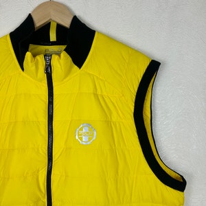 Puede incluir: Un chaleco acolchado amarillo con ribetes negros y un logotipo de Polo Sport plateado. El chaleco tiene una cremallera y un cuello alto.