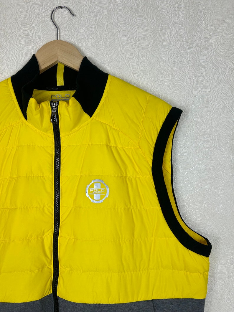 Puede incluir: Un chaleco acolchado amarillo con ribetes negros y un logotipo de Polo Sport plateado. El chaleco tiene una cremallera y un cuello alto.