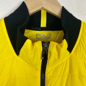 Puede incluir: Una chaqueta acolchada amarilla con cremallera negra y cuello negro. La chaqueta tiene el texto "POLO SPORT RALPH LAUREN" en la parte delantera.