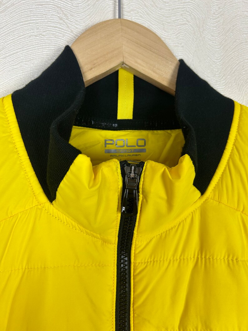Puede incluir: Una chaqueta acolchada amarilla con cremallera negra y cuello negro. La chaqueta tiene el texto "POLO SPORT RALPH LAUREN" en la parte delantera.