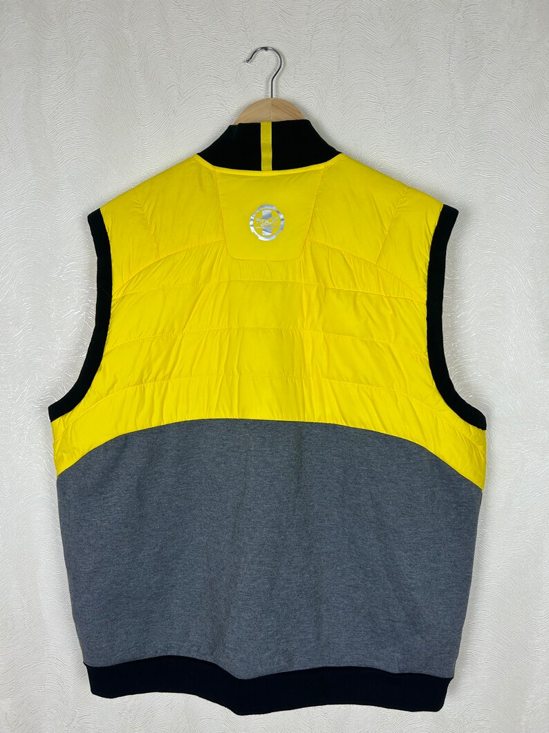 Puede incluir: Un chaleco sin mangas amarillo y gris con una cremallera negra y un cuello negro. El chaleco tiene un logotipo en la espalda que dice "Team".