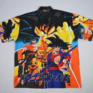 May include: Short-sleeved button-down shirt featuring a vibrant graphic of anime characters in shades of blue, orange, yellow, and black. The shirt has a black collar and a colorful design that covers the entire front and back.