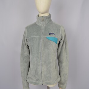May include: A gray fleece jacket with a zipper closure and a blue logo patch on the chest. The jacket has a stand-up collar and long sleeves.