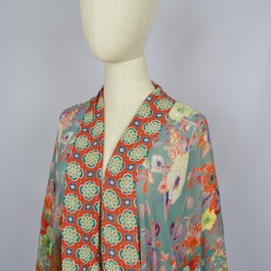 May include: A patterned kimono in teal, orange, and yellow, displayed on a mannequin. The kimono has a V-neck with an orange geometric panel. The sleeves and body are decorated with a vibrant floral design. The garment is made of a lightweight fabric.