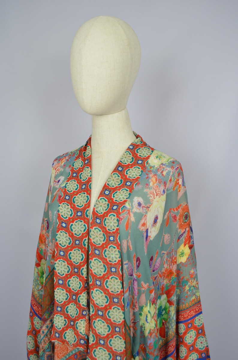 May include: A patterned kimono in teal, orange, and yellow, displayed on a mannequin. The kimono has a V-neck with an orange geometric panel. The sleeves and body are decorated with a vibrant floral design. The garment is made of a lightweight fabric.