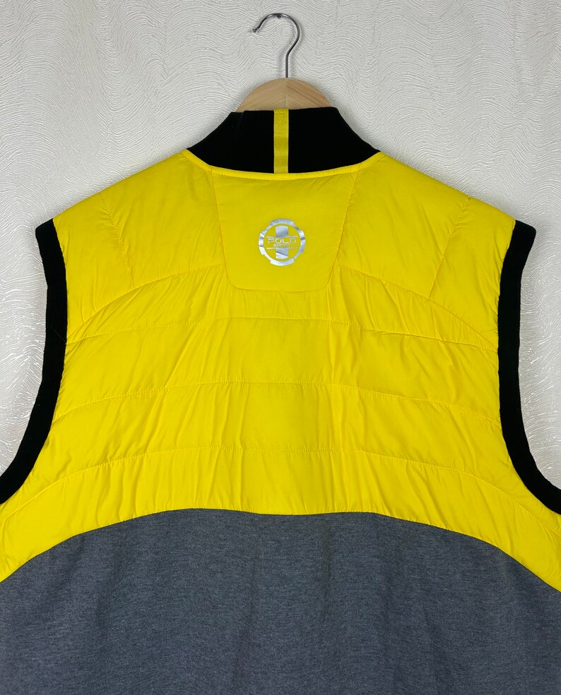 Puede incluir: Chaleco sin mangas amarillo y gris con cremallera negra y cuello negro. El chaleco tiene un logotipo en la espalda que dice "Polo Ralph Lauren".
