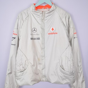 Può includere: Giacca grigio chiaro con fodera arancione e collo alto. La giacca presenta il logo Mercedes-Benz, il logo Vodafone e il logo Mobil 1. Ha due tasche anteriori ed è appesa a un appendiabiti azzurro chiaro.