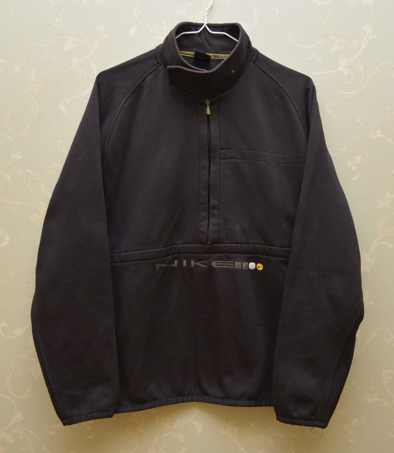 May include: A black Nike fleece jacket with a half-zip closure and a small pocket on the left chest. The Nike logo is embroidered in gray and yellow on the bottom front of the jacket.