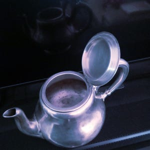 May include: A silver teapot with a lid that is open. The teapot is sitting on a black surface.