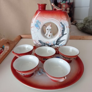 May include: A red and white ceramic sake set with a dragon and bird design. The set includes a bottle and six small cups.