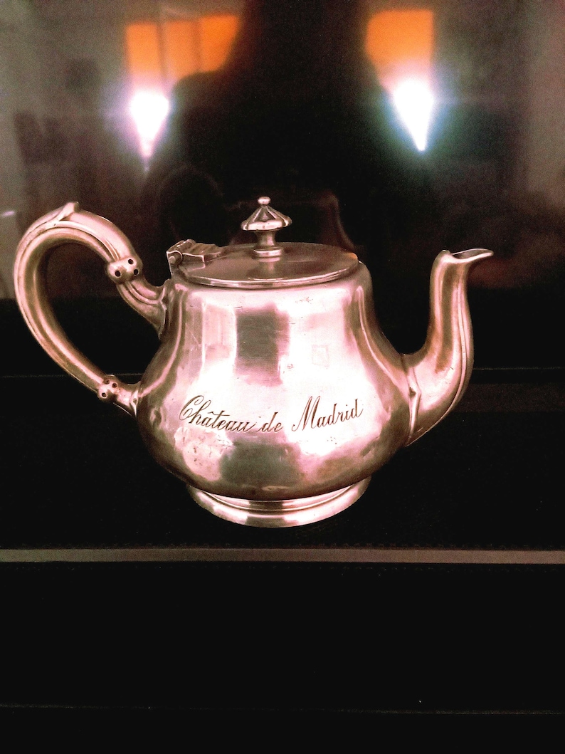 May include: A silver teapot with a round body and a curved spout. The teapot has an inscription that reads "Chateau de Madrid".