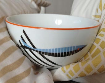 May include: A white ceramic bowl with an orange rim and a black and blue fish design. The bowl is sitting on a striped fabric.