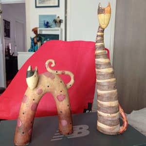 May include: Two ceramic cat figurines. One is brown and white with stripes and the other is brown with pink spots. Both have a whimsical, cartoonish style.