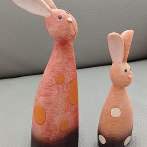 May include: Two ceramic bunny figurines, one tall and one short. The tall bunny is pink with orange polka dots and black accents. The short bunny is pink with white polka dots and black accents.