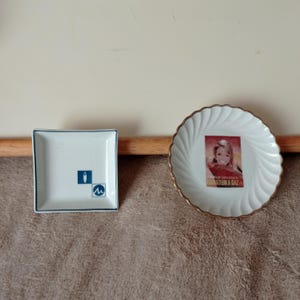 Puede incluir: Dos platos decorativos pequeños. Uno es cuadrado, blanco con borde azul y un logotipo azul y blanco. El otro es redondo, blanco con borde dorado y una imagen vintage de una mujer.