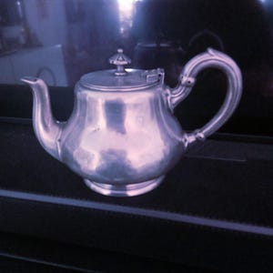 May include: A silver teapot with a rounded body and a lid with a knob on top. The teapot has a curved spout and a handle that curves back towards the body.