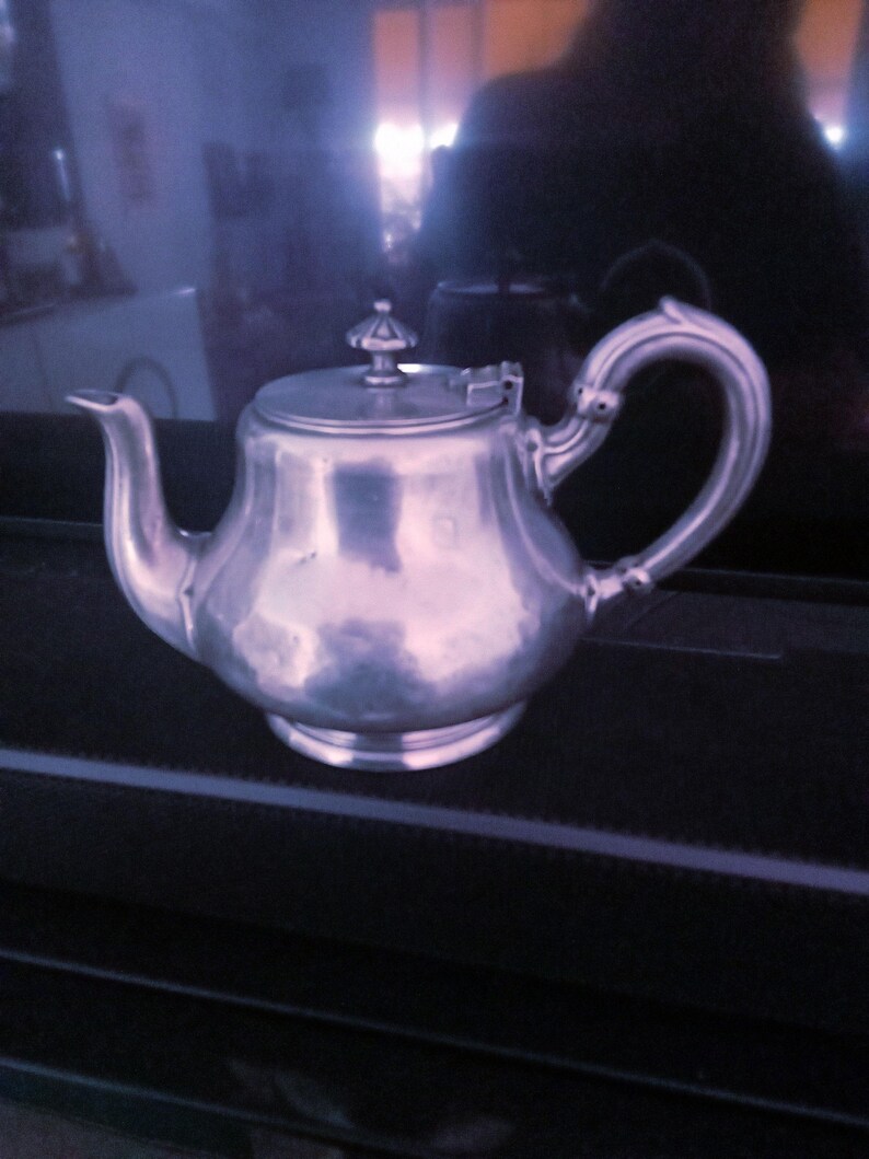 May include: A silver teapot with a rounded body and a lid with a knob on top. The teapot has a curved spout and a handle that curves back towards the body.