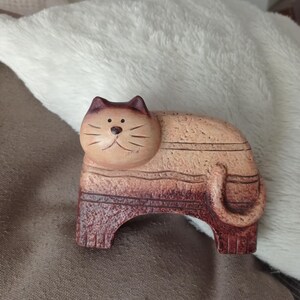 May include: A wooden cat ornament with a light brown body and dark brown stripes and details. The cat has a rounded head with a smiling face and a small tail. The ornament is on a brown surface.