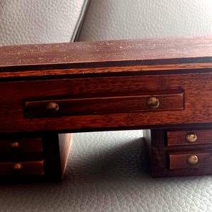 Peut inclure: Un bureau miniature en bois avec une finition brun foncé. Le bureau a un seul tiroir et trois tiroirs plus petits en dessous. Le bureau est en bois et a un design simple.