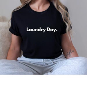 May include: Black t-shirt with the text "Laundry Day" in white. The shirt has short sleeves and a crew neck. The model is wearing gray sweatpants.