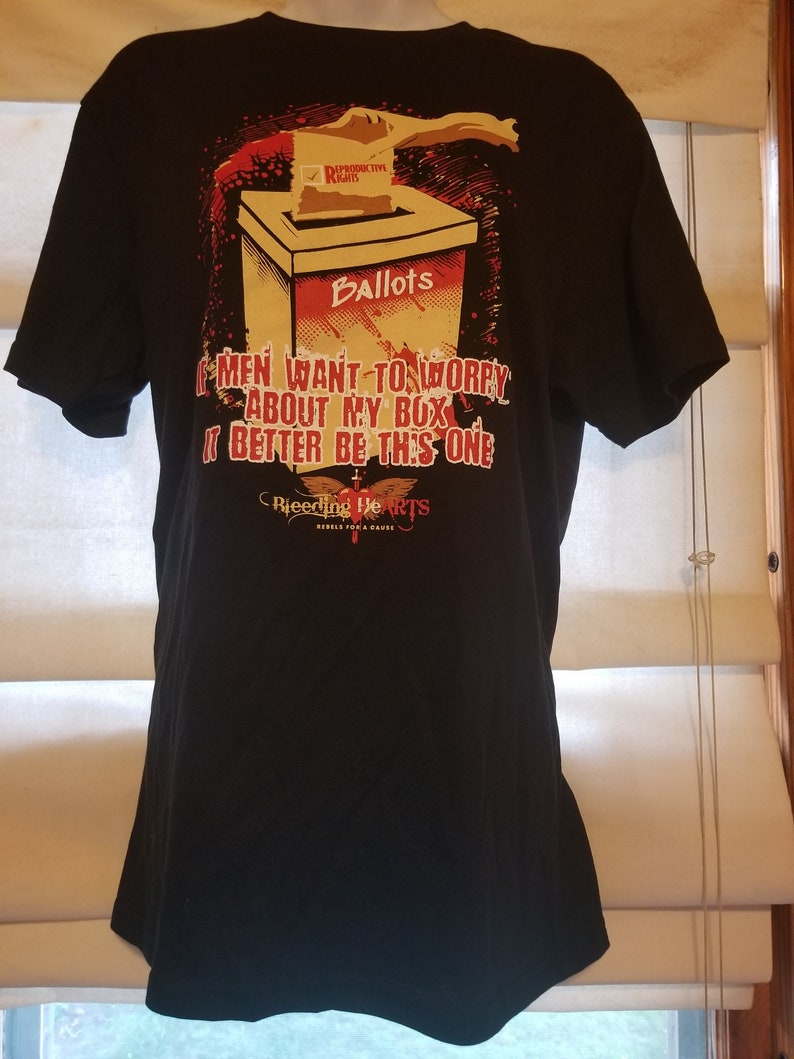 May include: Black t-shirt featuring a graphic of a hand dropping a ballot into a box labelled "Ballots". The ballot has a tick next to "Reproductive Rights". The text on the shirt reads "If men want to worry about my box, it better be this one." Below the graphic is the text "Bleeding Hearts Rebels for a Cause."