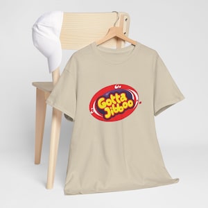 Könnte beinhalten: Beiges T-Shirt mit dem Text "Gotta Jibboo" in einem roten Oval-Design. Der Text ist gelb und lila mit weißen Akzenten. Eine weiße Baseballkappe hängt an einem Holzstuhl.