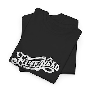 May include: Two black t-shirts are neatly folded and stacked. The top shirt displays the white text "Fluff Head" in a distressed, vintage-style font. The shirts are made of a soft material.