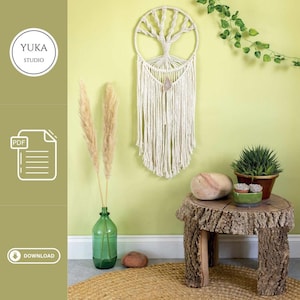 May include: A white macrame wall hanging in the shape of a tree with a light brown stone hanging from the bottom. The wall hanging is on a light green wall with a small wooden table with a light brown stone on top. The table is in front of a green plant in a brown pot.