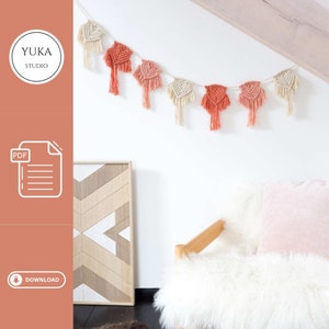 Puede incluir: Una descarga PDF para un patrón de macramé para colgar en la pared. El patrón presenta una guirnalda de nudos de macramé de color melocotón y crema. La guirnalda está colgada en una pared blanca sobre un sofá blanco con un cojín de melocotón.