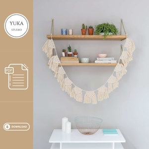 Puede incluir: Una guirnalda de macramé blanca con borlas cuelga de dos estantes de madera. Los estantes están decorados con plantas en macetas, velas y libros. La guirnalda es un proyecto de bricolaje y la descarga PDF está disponible.
